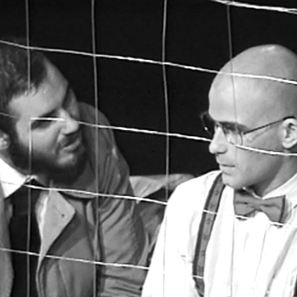 Two men in conversation behind barbed wire. The image is in black and white.